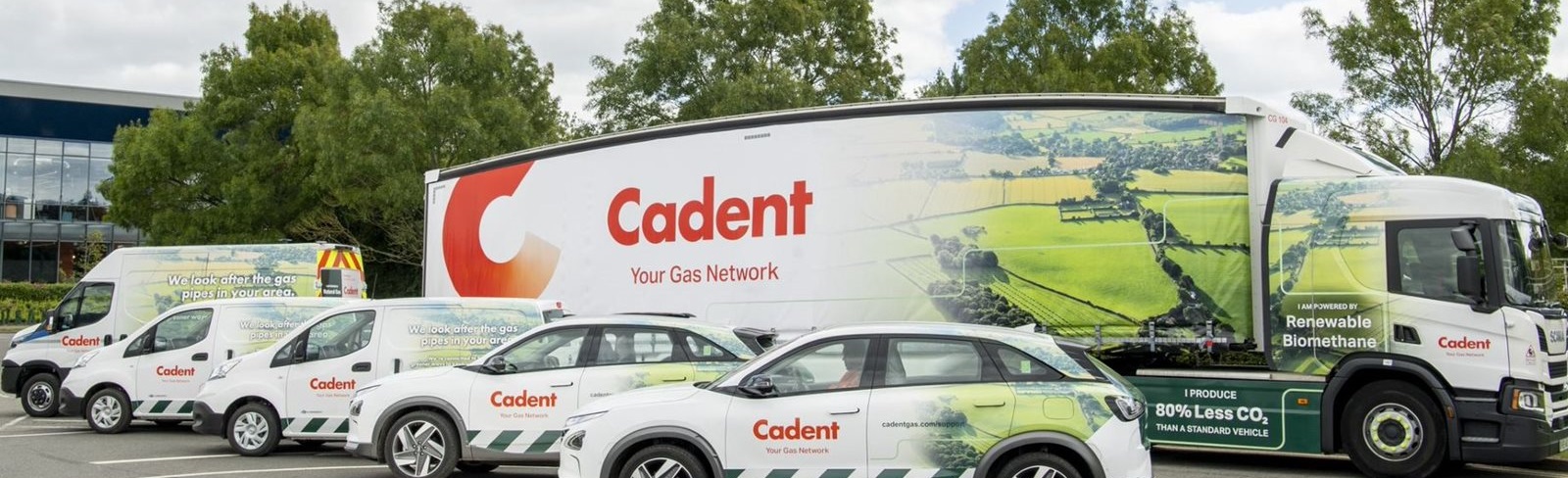 Cadent-branded fleet of vehicles including a large truck and several cars parked in a lot, featuring renewable biomethane and low CO₂ messaging.