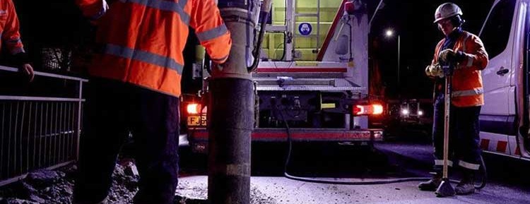 Construction workers in reflective orange jackets operate heavy machinery near a truck at night, highlighting an active work zone with focused determination.