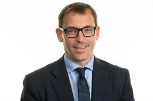 A man in a suit with glasses smiles confidently at the camera against a plain white background, conveying professionalism and approachability.