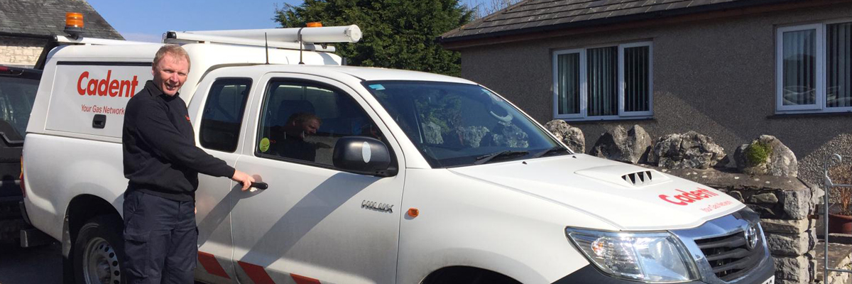 Man in a black shirt stands beside a white utility truck with "Cadent" logo, parked in front of a stone house. The scene is sunny and cheerful.