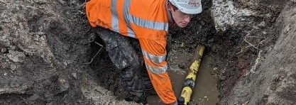 A construction worker in an orange safety jacket and helmet repairs a yellow pipe in a muddy trench. The scene conveys a sense of focus and effort.