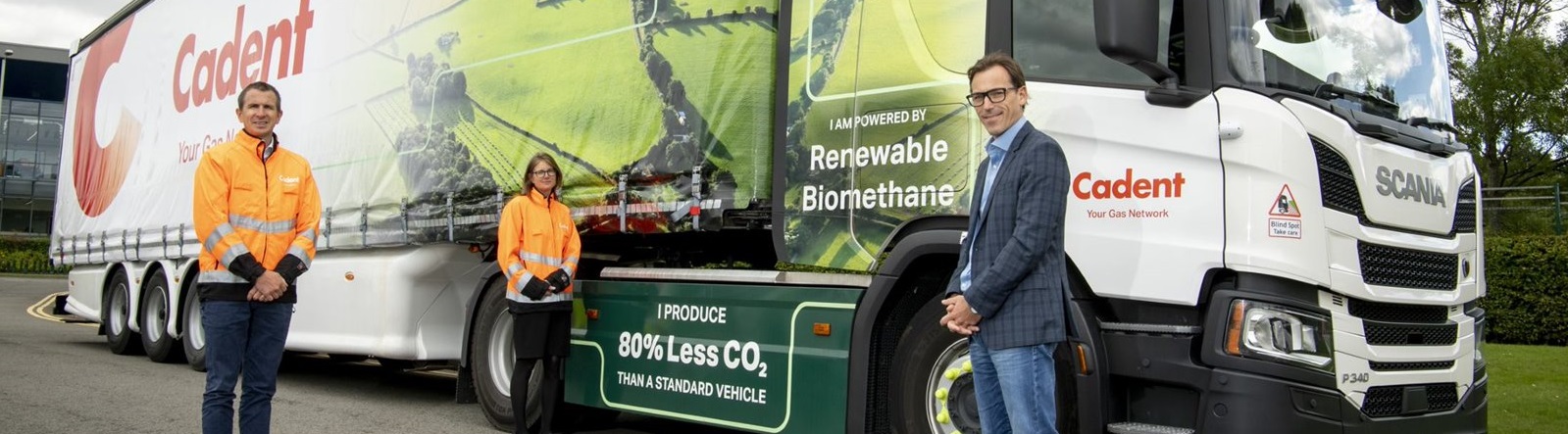 Three people stand in front of a large truck with 'Cadent' branding. The truck announces it runs on renewable biomethane, reducing CO2 emissions by 80%. The mood is positive and eco-friendly.