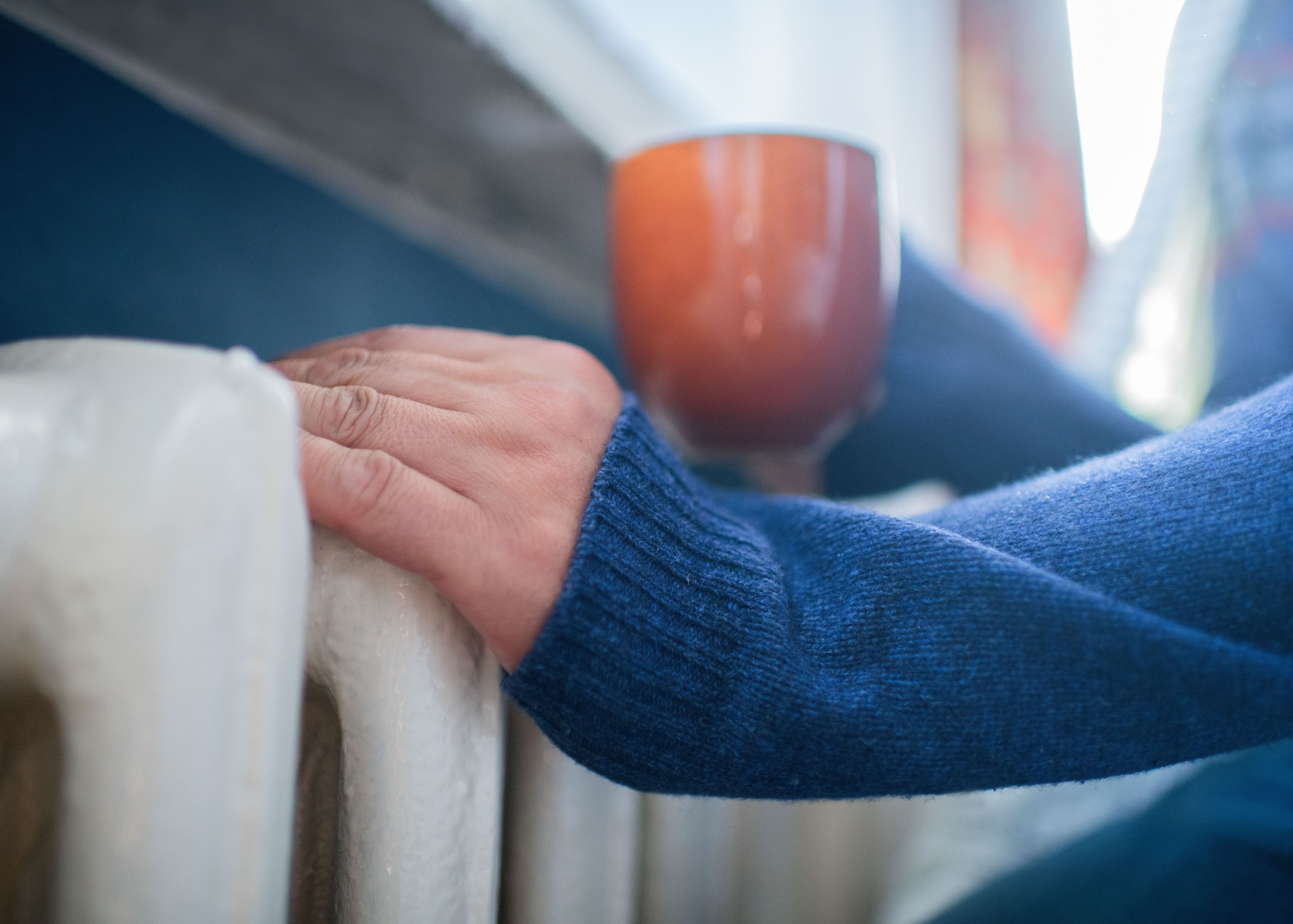 Man in warm sweater with hot tea near radiator in winter. 