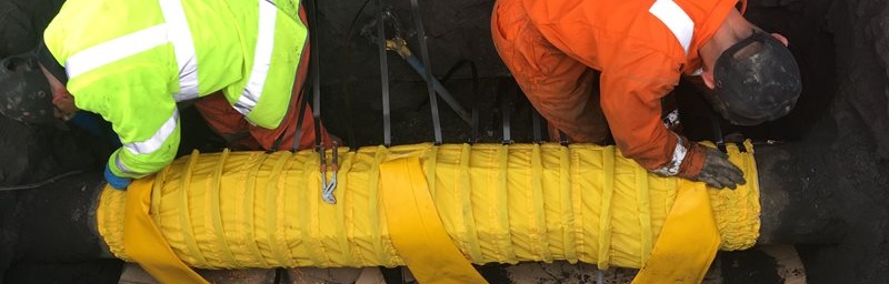 Two workers in high-visibility clothing, one in yellow and one in orange, are in a trench securing a large pipe wrapped in yellow insulation, conveying teamwork.