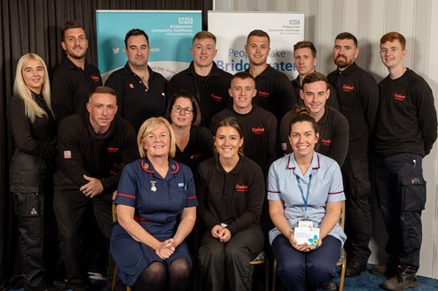 01_e A group of fifteen people, including men and women in uniform, pose together indoors. They appear positive and professional, with two seated women in front.
