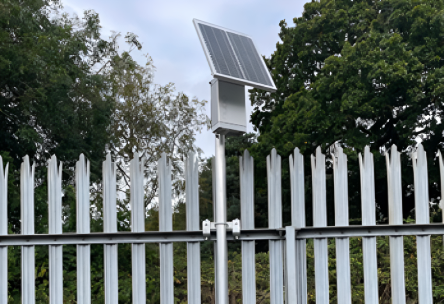 monitoring unit on a fence A small solar panel mounted on top of a metal box fixed to a tall metal fence, surrounded by trees in the background.