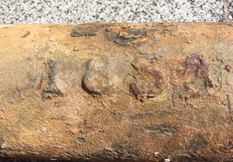 Close-up image of a rusted metal pipe or cylinder with the engraved marking “1859” partially visible on its surface, showing signs of corrosion and aging.