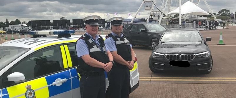 Two uniformed police officers stand next to a patrol car on a cloudy day. Behind them is a large Ferris wheel and several parked cars, creating a busy, watchful scene.