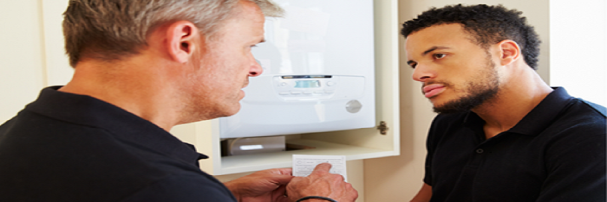 Two men are inspecting a wall-mounted boiler inside a cabinet. The older man points at instructions while the younger man listens attentively.