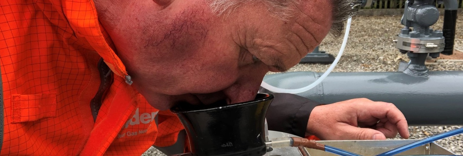 A person in an orange safety jacket inspects a metal gas monitoring device outdoors, focusing intently. Pipes and gauges are visible in the background.