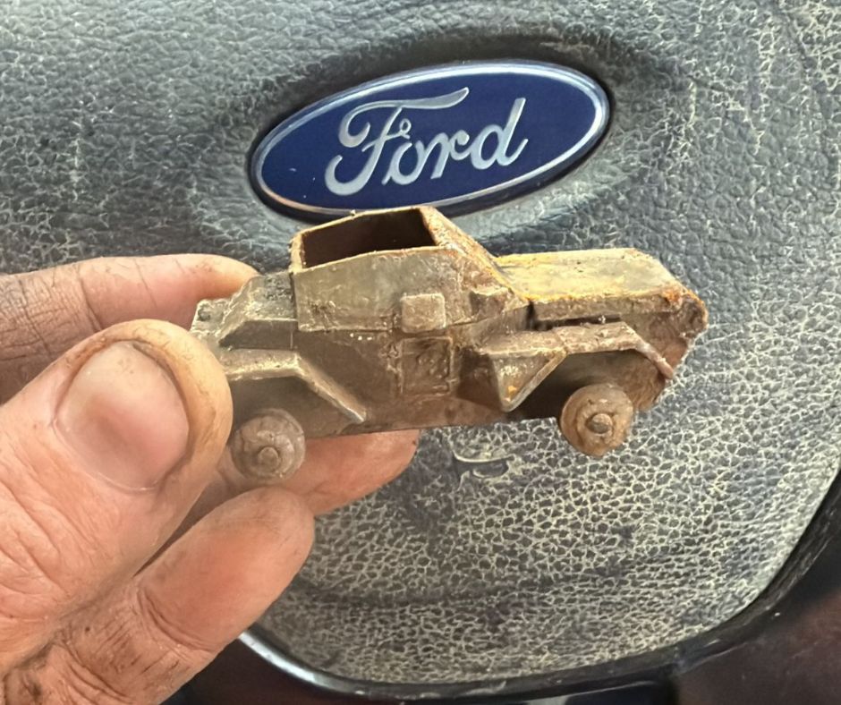 A small rusted metal toy car found inside a gas pipe, held up against a vehicle steering wheel.