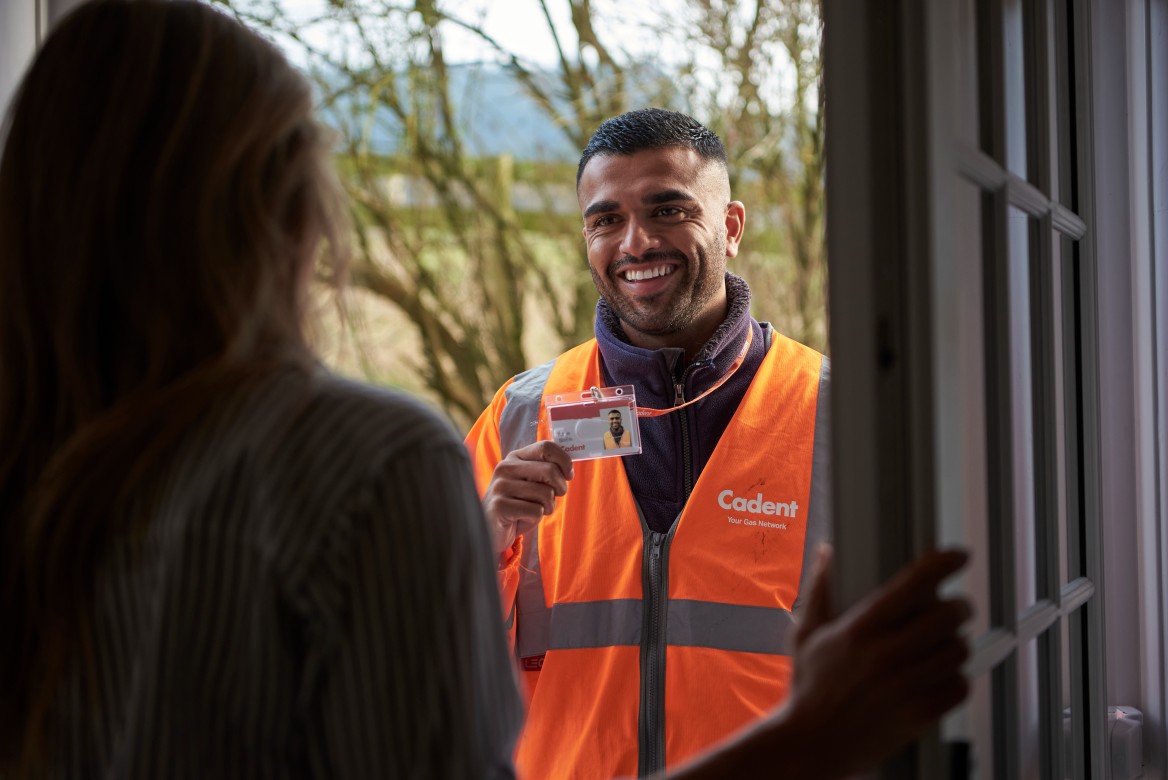 A smiling utility worker in a high-visibility orange vest holds an ID badge at a doorstep. Trees are visible in the background.