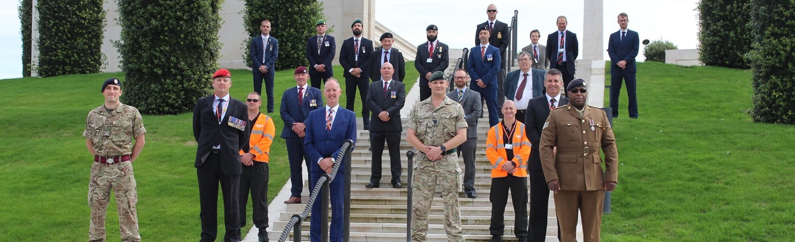 A group of people, including military personnel in camouflage and uniforms, stand solemnly on steps in front of tall trees and a memorial, conveying respect.