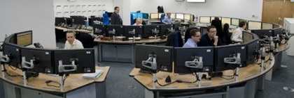 A spacious control room with curved desks, multiple computer monitors, and focused staff. The atmosphere is busy and professional, displaying teamwork and technology.