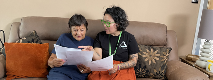Two people sit on a couch, reviewing documents together. The person on the right points at the paper, suggesting collaboration and focus.