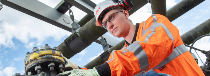 Industrial worker in high-visibility orange safety gear and hard hat inspecting a bolted pipeline connection at an outdoor industrial facility.