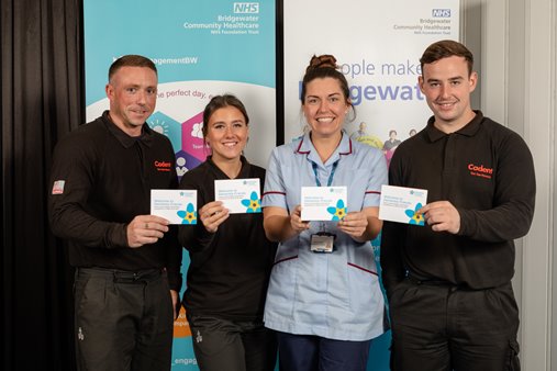 Four individuals stand smiling, each holding a certificate. Three wear black uniforms, one is in a nurse's uniform. Background features NHS banners.