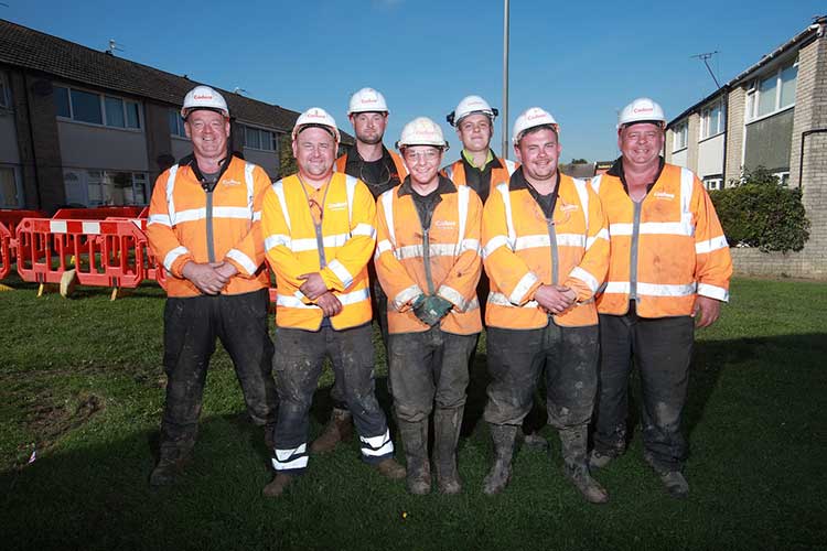 Seven construction workers stand on a grassy area in front of houses, wearing orange safety jackets and white helmets, smiling in the sunlight_image