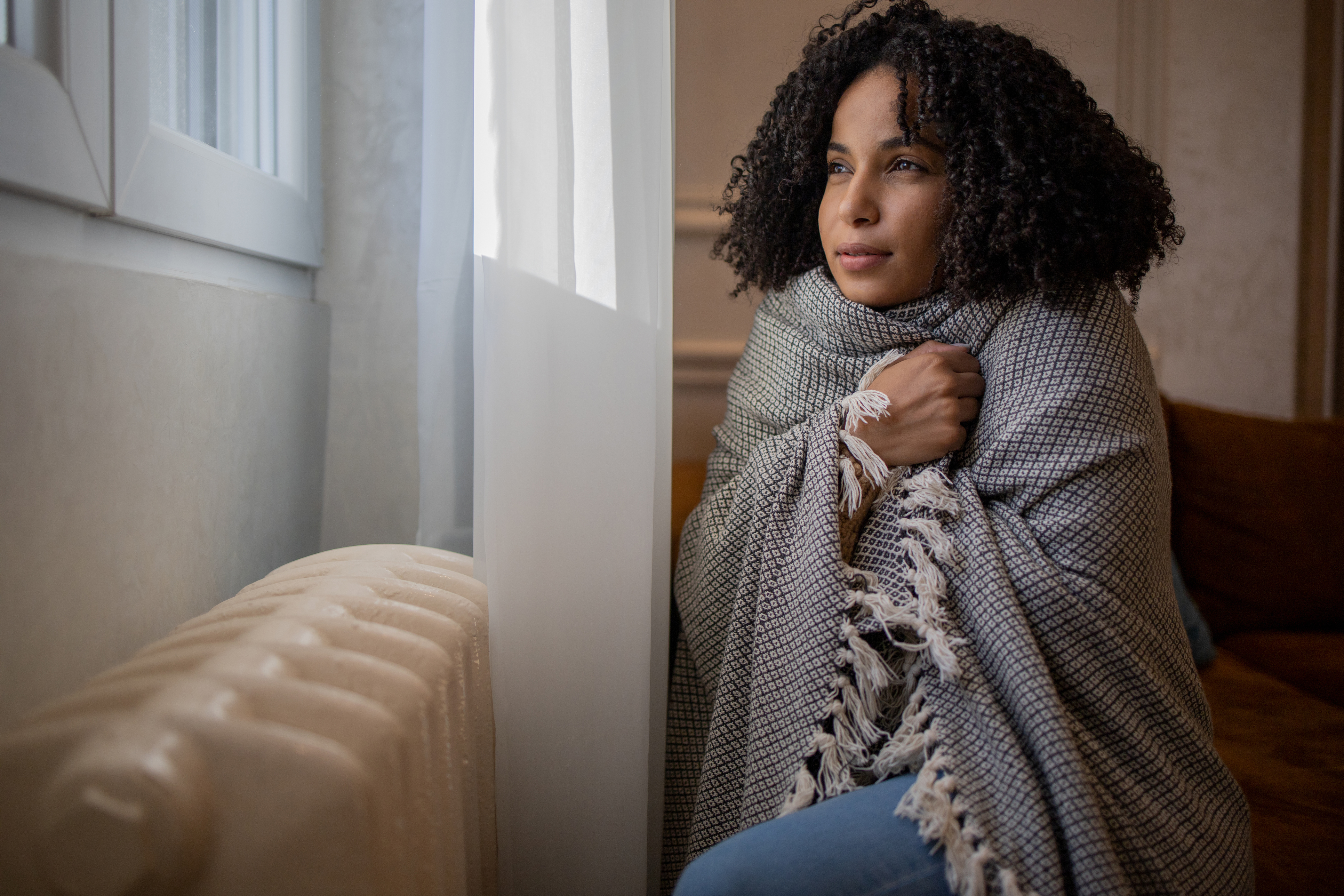 A woman with curly hair looks thoughtfully out the window, wrapped in a cozy blanket. She sits beside a radiator, conveying warmth and comfort.