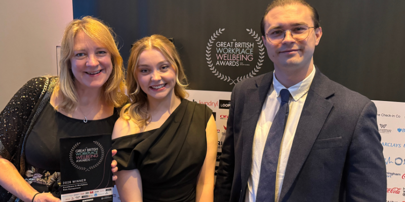 A group of three people pose together at an event, standing in front of a Great British Workplace Wellbeing Awards backdrop. One person holds an award plaque as they gather for a photo.
