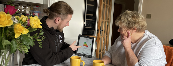 A man shows an elderly woman a tablet with a colorful chart. They are seated at a table with yellow mugs and a flower vase, discussing attentively.