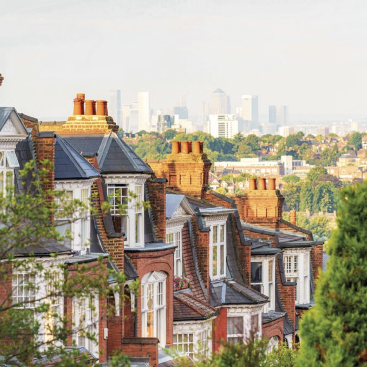 Row of charming red-brick houses with slate roofs under a clear sky. In the background, a hazy city skyline is visible, surrounded by greenery.