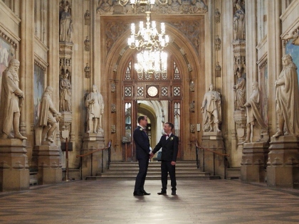 Two people in formal suits shaking hands in a grand hall with ornate architecture, statues, and chandeliers.