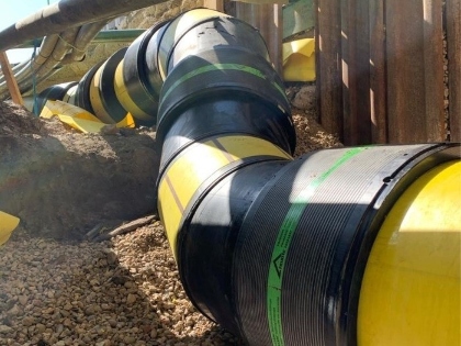 Curved yellow gas pipes with black protective joints installed in a trench surrounded by gravel and metal panels