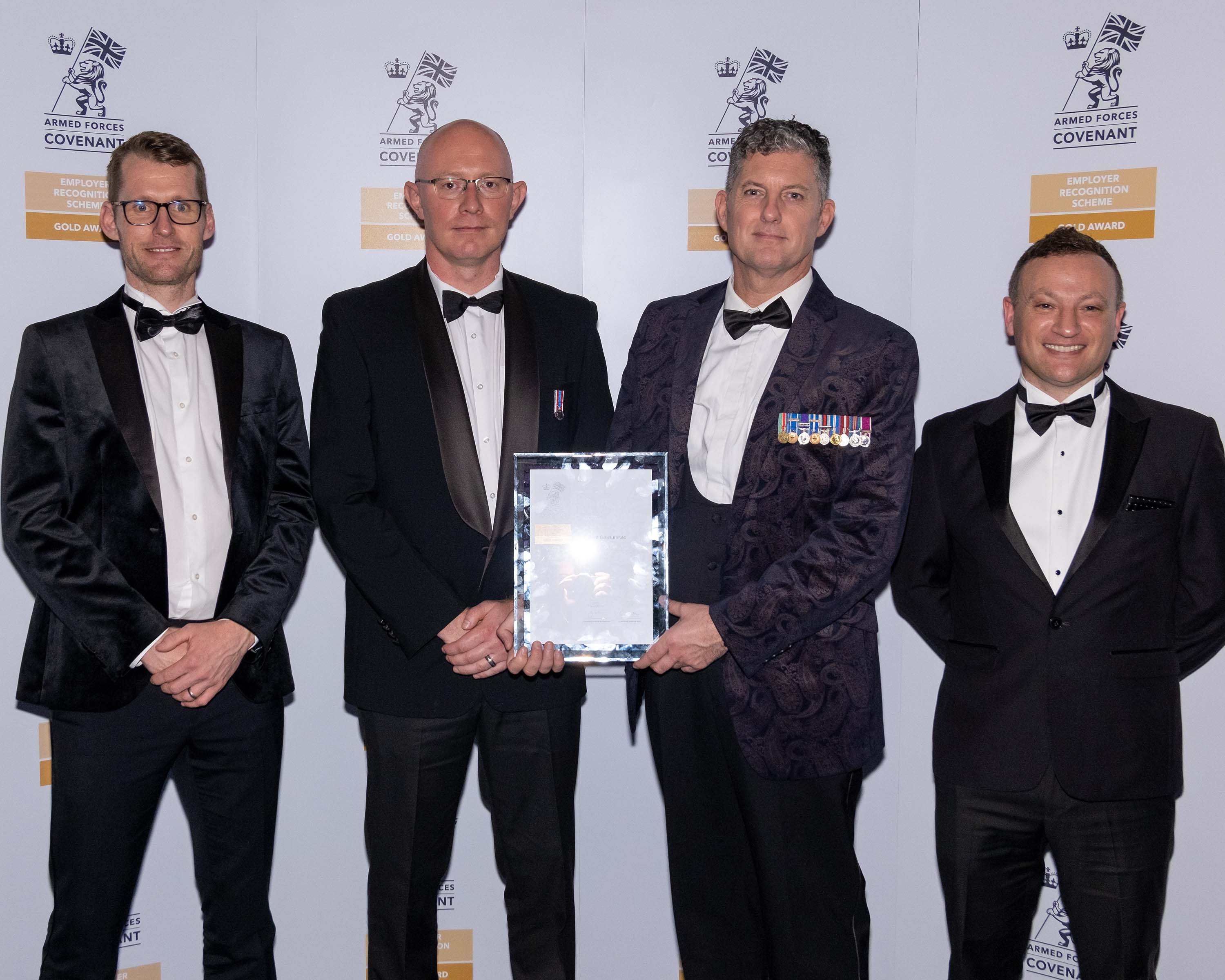 Four people in tuxedos posing with an award in front of a branded backdrop