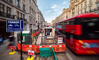 Busy city street with construction barriers and equipment in the center, a red double-decker bus passing by, and a blue taxi/bicycle lane sign on the left.