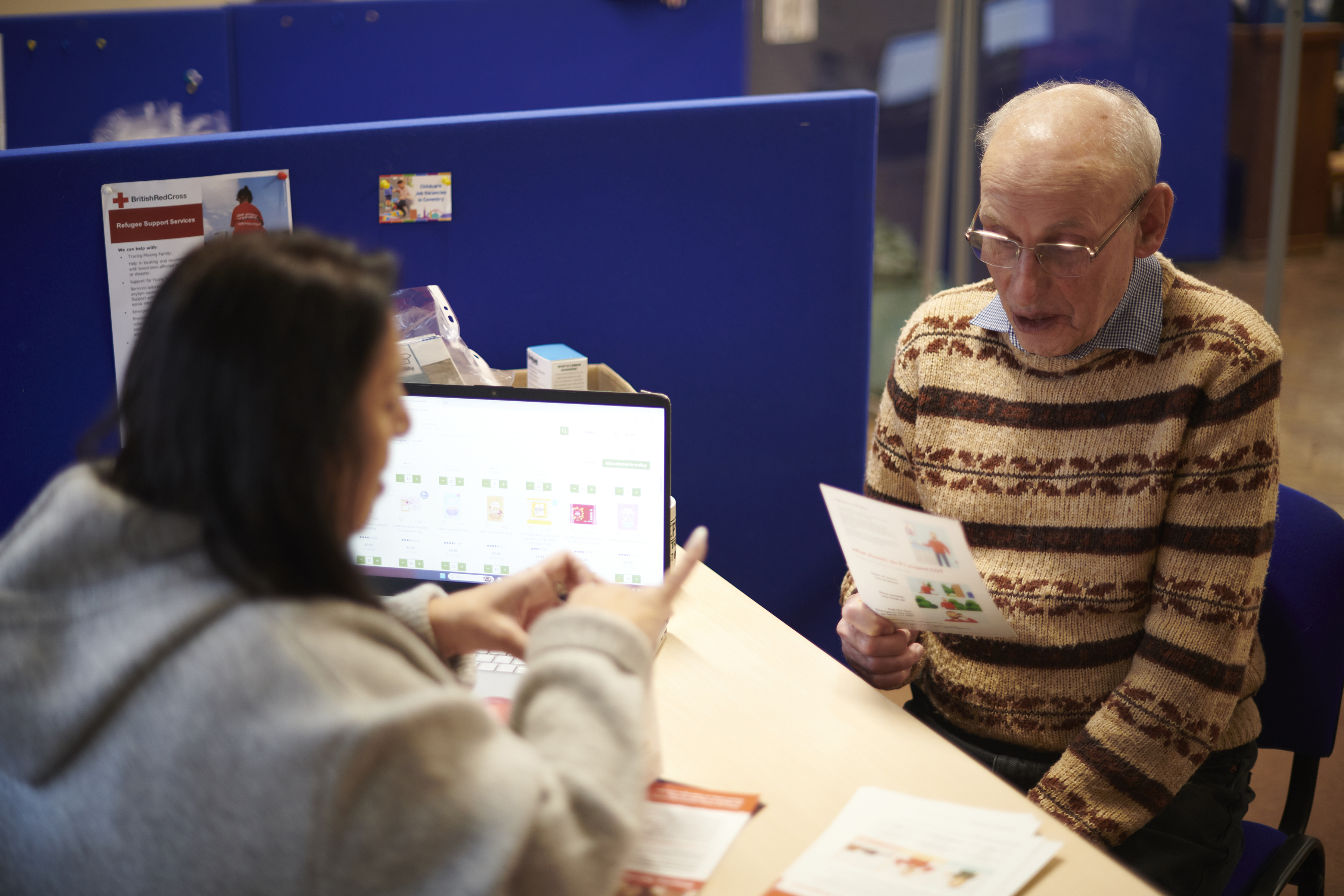 An older man in a patterned sweater discusses paperwork with a staff member at a computer in a blue-partitioned service office, calmly.