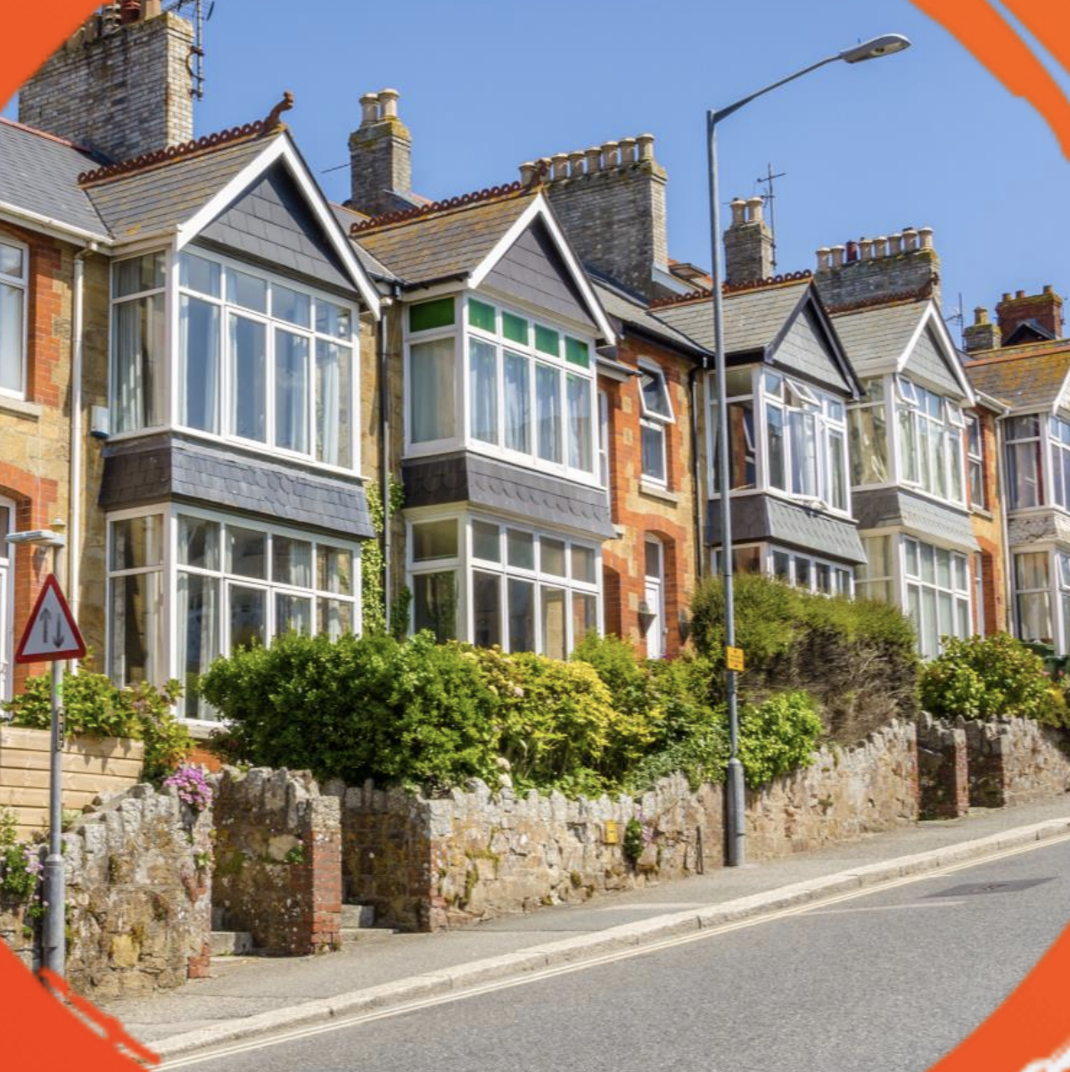 Row of charming Victorian houses with bay windows, stone walls, and lush greenery on a sunny day. The street is sloped, enhancing the quaint setting.