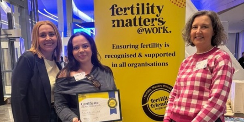 Three women standing indoors in front of a yellow ‘Fertility Matters at Work’ banner. The woman in the center is holding a Certificate of Accreditation. All three are smiling at the camera.