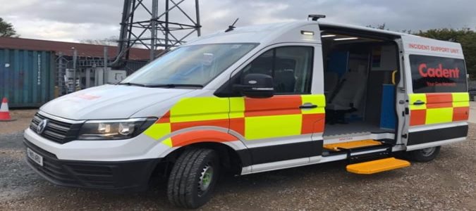 White incident support van with red and yellow reflective stripes is parked with its side door open, displaying a yellow step. Overcast sky in the background.