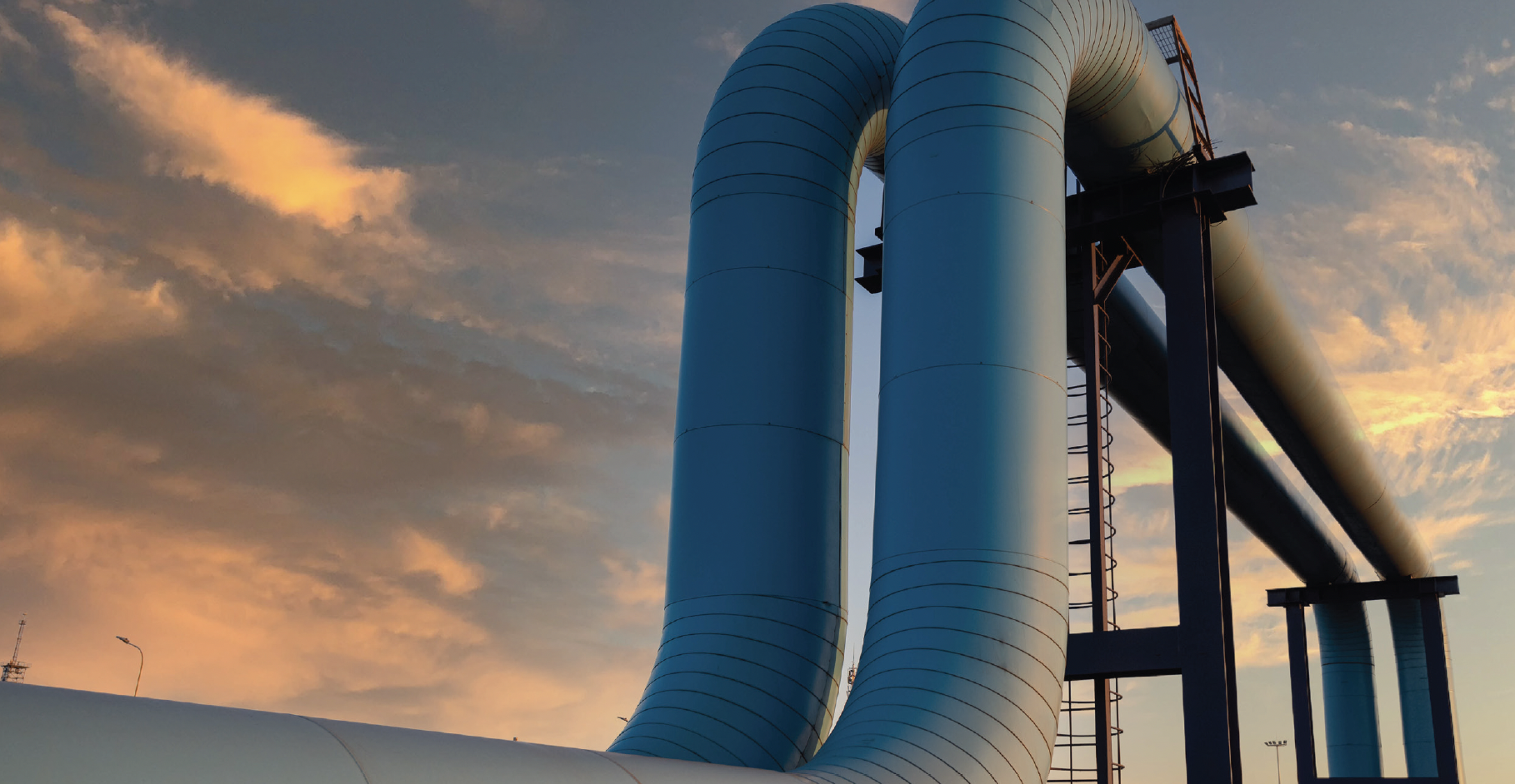 Large blue industrial pipes curve upward against a vibrant sunset sky, creating a striking contrast. The scene conveys a sense of scale and industry.