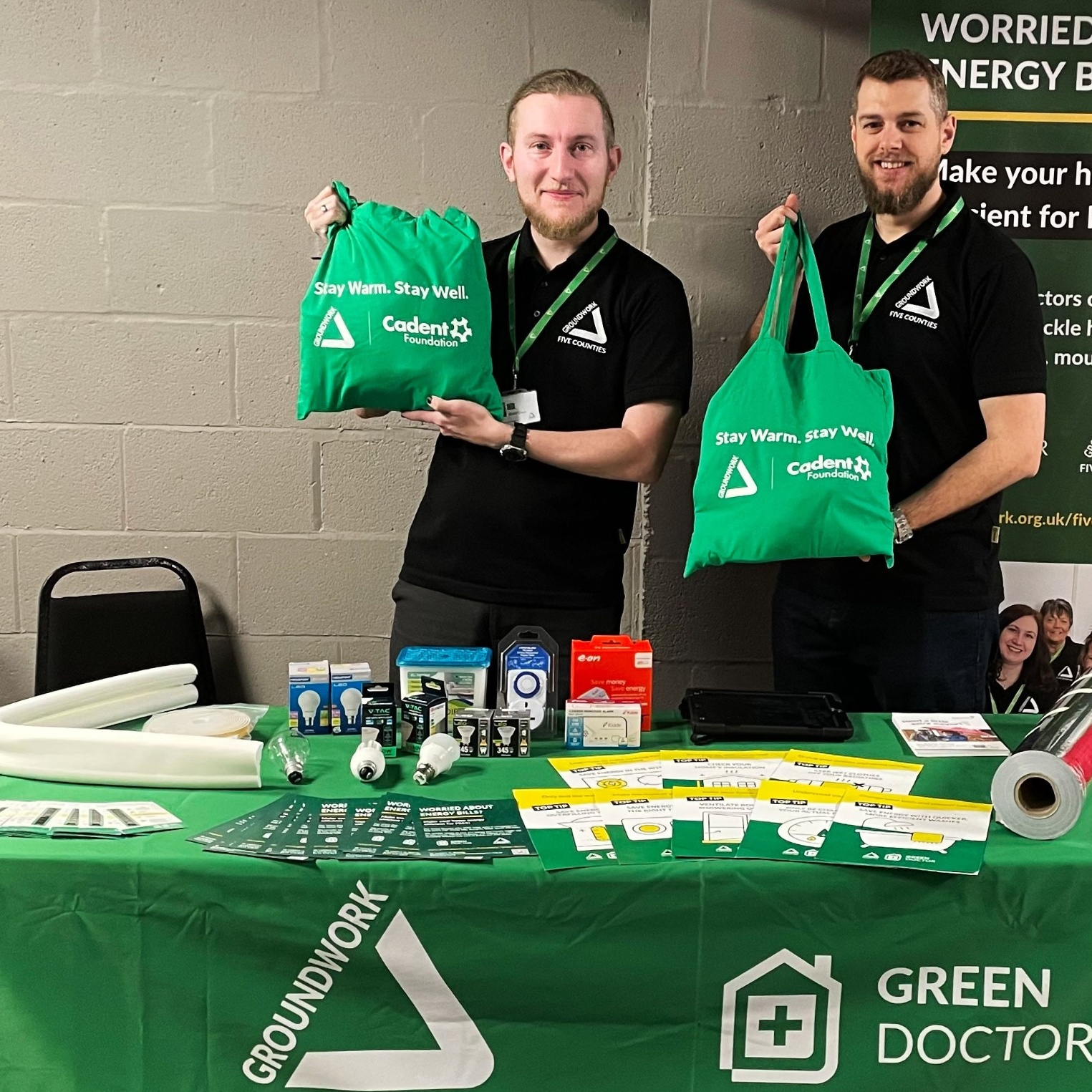 Two smiling individuals hold green bags with 'Stay Warm, Stay Well' text at a booth. The table displays energy-saving items. Warm, informative atmosphere.