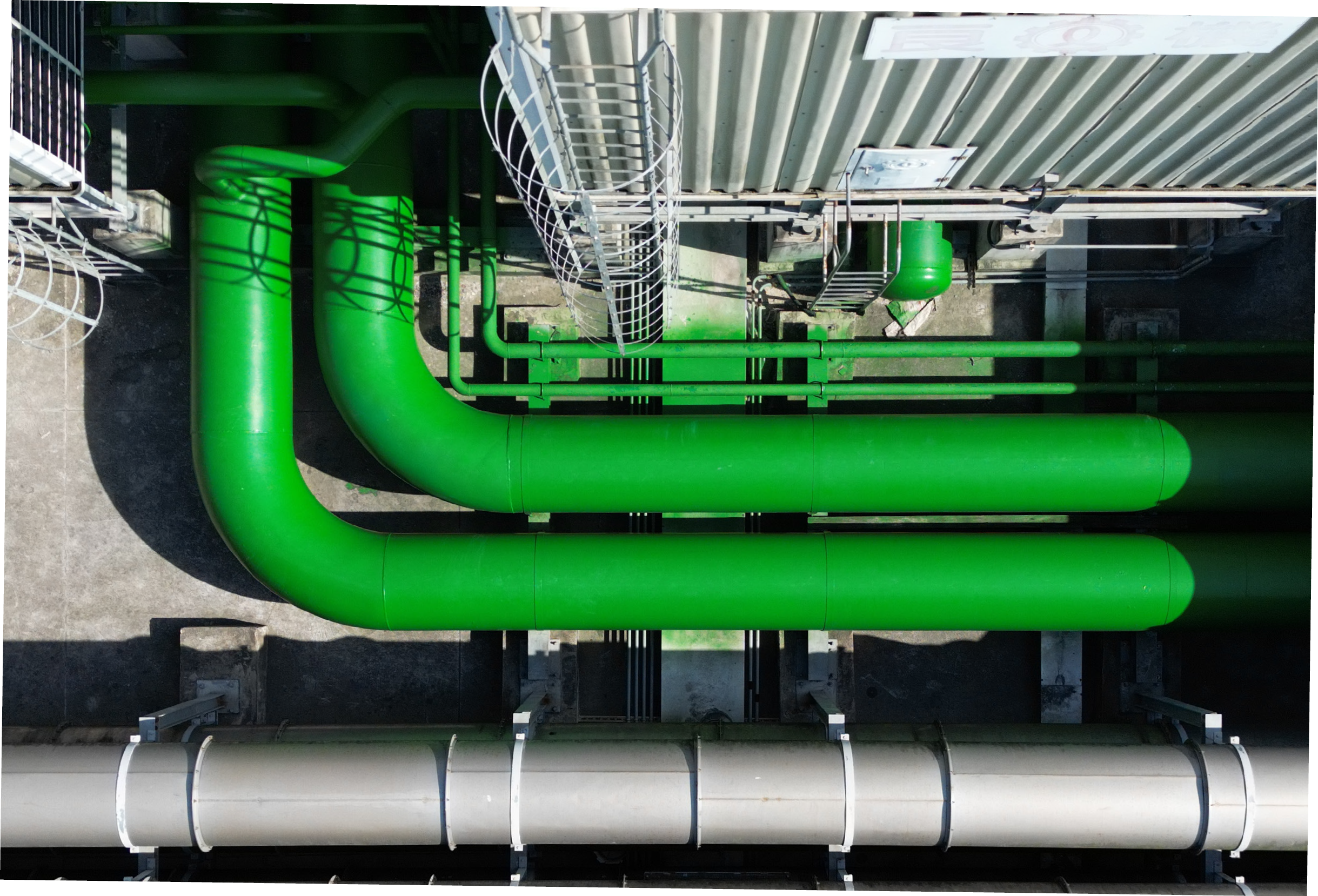 Aerial view of bright green industrial pipes running parallel on a concrete surface. A ladder and railing are visible, creating a symmetrical pattern.