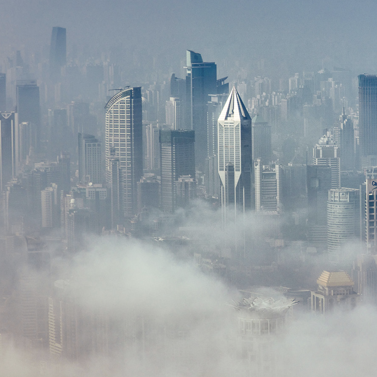 A dense fog shrouds a cityscape, with skyscrapers piercing through the mist. The tallest, a sharp, angular building, contrasts against the hazy skyline. Calm and mysterious.