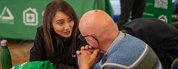A woman with long brown hair attentively listens and talks to an elderly man in a blue sweater. They sit at a table with a green background, conveying a supportive atmosphere.