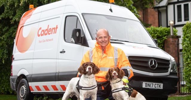 A man in an orange safety jacket kneels with two dogs in front of a white van labeled 'Cadent' The scene is outdoors, conveying readiness and teamwork.