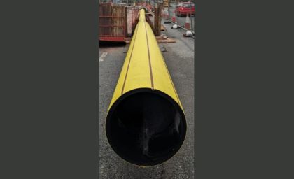 A large yellow pipe lies horizontally on a construction site with traffic cones and barriers. The scene conveys a sense of urban development and industrial work.