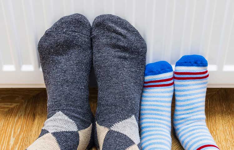 Two pairs of feet in cozy socks, one gray and argyle, the other blue with stripes, rest on a warm radiator. The scene conveys warmth and comfort.