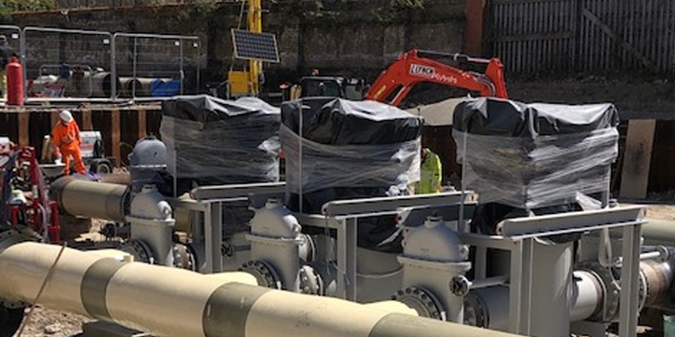 Industrial gas pipes and valves wrapped in protective plastic at a construction site, with workers and machinery in the background.