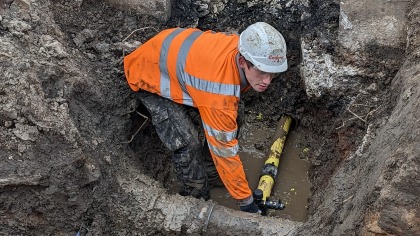 A construction worker in an orange safety jacket and helmet repairs a yellow pipe in a muddy trench. The scene conveys a sense of focus and effort.