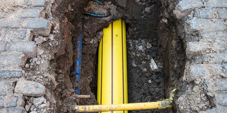 Yellow gas pipes exposed in a dug trench surrounded by soil and cobblestone pavement.