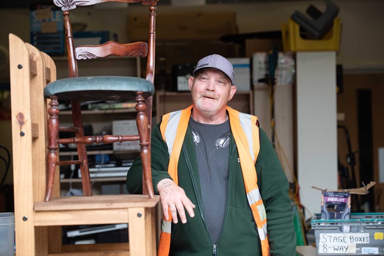 A man in a workshop leans on stacked wooden chairs, wearing a gray cap and orange vest. The setting has tools and boxes, conveying a casual, industrious mood.