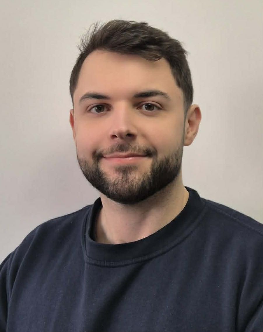 Portrait of a young man with dark hair and a short beard wearing a navy shirt, facing the camera against a plain background with a calm smile.