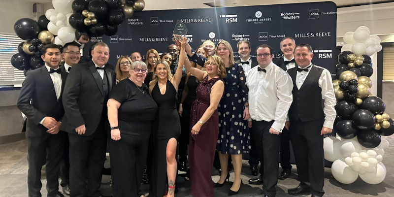 A large group of people dressed in formal evening wear posing together at an awards event. Several individuals in the center are holding up a trophy. Behind them is a branded backdrop and black, white, and gold balloon displays.