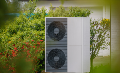 Outdoor air-source heat pump unit installed beside a house, featuring two large black circular fans on a grey panel, surrounded by green plants and shrubs in a garden setting.